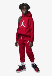 Sudadera de forro polar roja con logo blanco, pantalones de chándal a juego y zapatillas negras con acentos rojos. Mochila de camuflaje adicional colgando de un hombro.