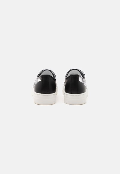 Une paire de baskets basses noires avec des semelles blanches vues de dos sur un fond clair uni.