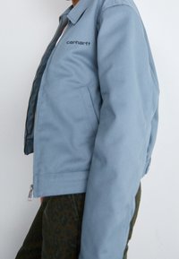 Giacca azzurro chiaro con chiusura a zip, colletto aperto e tasche laterali. Presenta un logo nero a contrasto sul lato sinistro del petto.