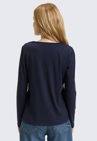 Granatowa bluzka z długim rękawem o okrągłym dekolcie, gładkiej fakturze i dopasowanym kroju, zestawiona z niebieskimi dżinsami, widziana od tyłu.