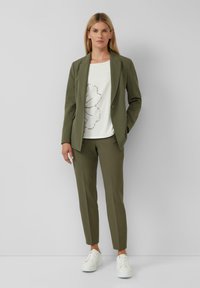Olijfgroen pak met slim-fit broek en blazer; wit shirt met een bladontwerp. Gecombineerd met witte sneakers. Soepele textuur.