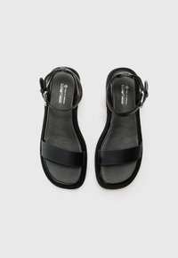 Sandales véganes noires avec une semelle plate, deux larges brides sur le dessus du pied et une bride ajustable à la cheville dotée d'une boucle en argent.