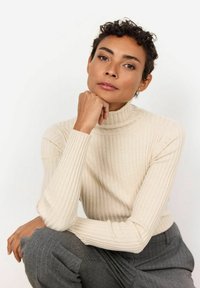 Beige, ribbed turtleneck sweater with long sleeves, paired with gray pinstriped trousers. Model rests chin on hand, displaying a neutral expression.