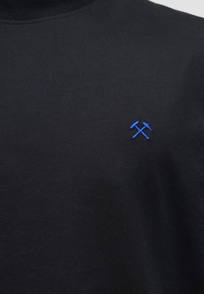 Μαύρο ύφασμα με μικρό μπλε κεντητό σχέδιο που απεικονίζει σφυρί και λοστό σταυρωμένα στην αριστερή πλευρά του στήθους.