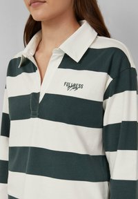 Vrouw in een rugbyshirt met kraag met brede donkergroene en witte horizontale strepen en de tekst "FULLNESS" geborduurd op de borst.