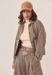 Mujer con una gorra beige, un suéter de punto crema de cuello alto y un conjunto a cuadros a juego de chaqueta y pantalones, complementado con un cinturón ancho topo con detalles metálicos.