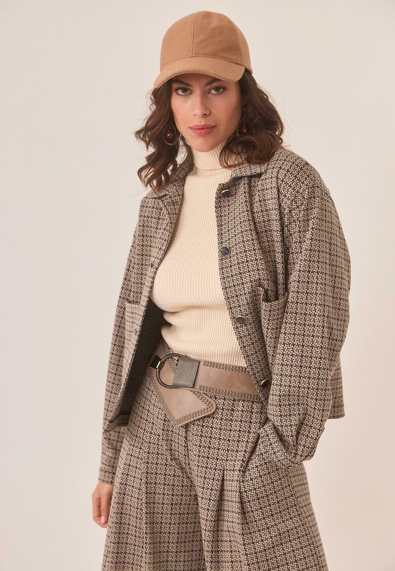 Mujer con una gorra beige, un suéter de punto crema de cuello alto y un conjunto a cuadros a juego de chaqueta y pantalones, complementado con un cinturón ancho topo con detalles metálicos.