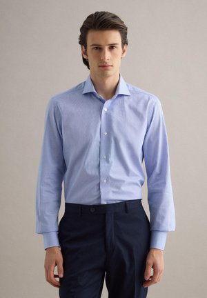 Giovane uomo con capelli scuri indossa una camicia elegante azzurro chiaro con bottoni e pantaloni blu scuro, in piedi davanti a uno sfondo neutro.