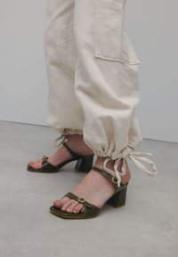 Groene leren blockheel sandalen met open tenen, dunne bandjes en ronde gouden accenten, gedragen met beige elastische broekbanden.