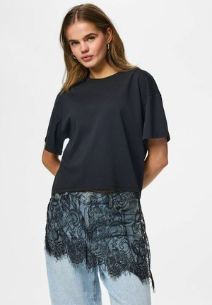 Ung kvinde med mellemlangt hår iført en løs sort T-shirt og lyseblå jeans med sort blonde foran.