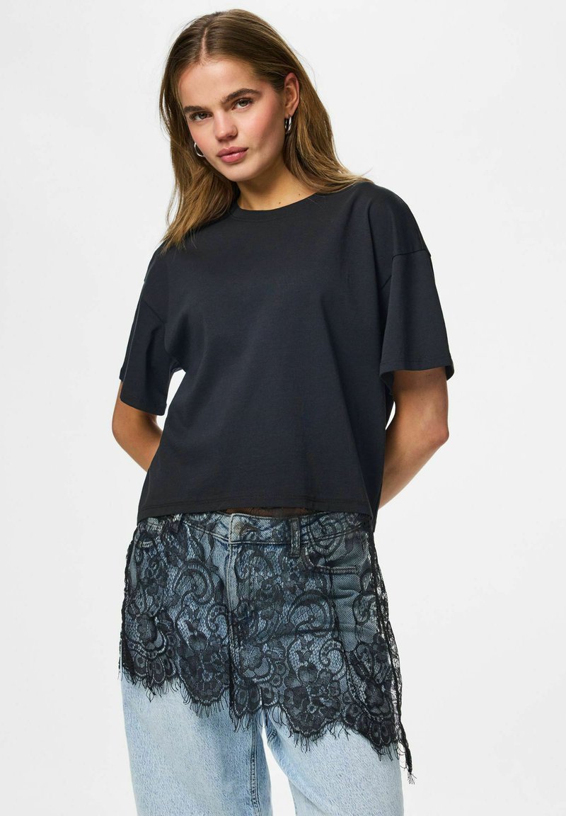 Jeune femme aux cheveux mi-longs portant un t-shirt noir ample et un jean bleu clair avec un empiècement en dentelle noire à l'avant.