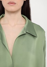 Femme portant une chemise en satin vert clair avec un col ouvert, dévoilant le décolleté ainsi qu'une partie de ses lèvres et de son menton, avec de longs cheveux foncés.