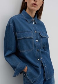 Camicia in denim blu scuro con bottoni a pressione in argento, dotata di due tasche sul petto e maniche lunghe con risvolti.