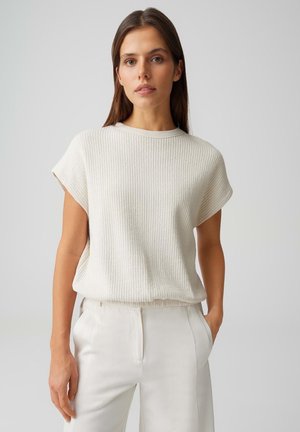 Vrouw draagt een crèmekleurige, geribbelde korte mouw gebreide top, ingestopt in een hooggetailleerde crèmekleurige broek, staand tegen een effen lichte achtergrond.