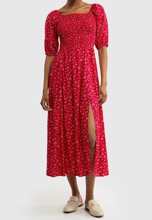 Nainen pukeutuneena punaiseen kukalliseen midimittaiseen mekkoon, jossa on puffy-hihat ja reiden korkeudella oleva halkio, yhdistettynä beigeihin liukuvastaisiin loafer-kenkiin.