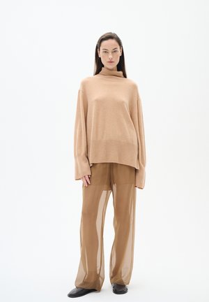 Bež turtleneck pulover s rebrastim rubom, uparen s hlačama od prozirnog smeđeg materijala visokog struka. Model nosi crne slip-on cipele.