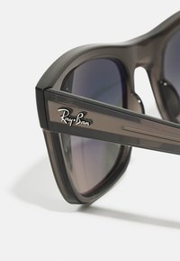 Ochelari de soare Ray-Ban cu un cadru gros negru, lentile gri cu gradient și un accent metalic cu logo pe braț. Textură mată cu linii de design subtile.