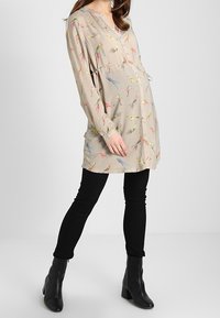 Umstandstunika aus beigem Stoff mit Vögel-Druck, ausgestattet mit einem V-Ausschnitt, langen Ärmeln und einer Empire-Taille, kombiniert mit schwarzen Skinny-Hosen.