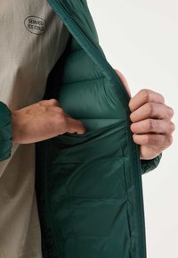 Mains ouvrant la veste matelassée verte pour montrer la poche intérieure, avec une chemise beige visible en dessous portant l'inscription "SERVED ICE COLD".