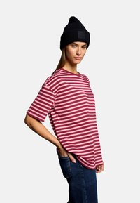 Red and pink striped t-shirt with short sleeves, paired with a black knitted beanie, model stands with one hand in denim pocket.