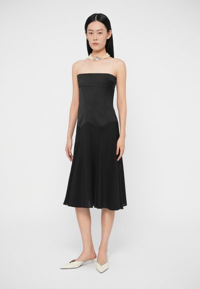 Žena v čiernych midi šatách bez ramienok, doplnených bielymi nízkymi lodičkami s ostrou špičkou a vrstveným perlovým náhrdelníkom so strieborným príveskom.