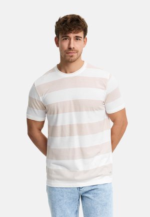 Ung mand med kort krøllet hår iført en hvid og beige stribet kortærmet skjorte og lyseblå jeans, stående med hænderne bag ryggen.