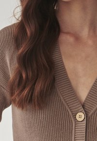 Bruine gebreide cardigan met een V-hals, geribbelde structuur en een houten knoopsluiting aan de voorkant. Zachte stof met verticale lijnen.