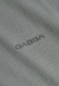Siva tkanina z fino teksturo, ki vsebuje utisnjen logo "GABBA" v temnejši sivi barvi, osredotočen na material.