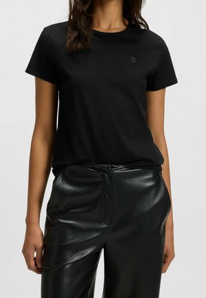 Kobieta w jednolitej czarnej koszulce z krótkim rękawem i czarnych skórzanych spodniach z wysokim stanem, stojąca na jednolitym tle.