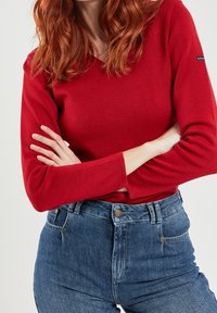 Rotes langärmliges Oberteil mit V-Ausschnitt und gerippter Textur, kombiniert mit hoch taillierten blauen Denim-Jeans mit Knopfverschluss und Fronttaschen.