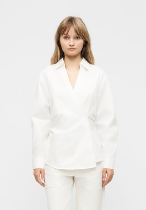 Sieviete ar gaiši brūniem matiem, valkājot baltu garām piedurknēm ar apkakli blūzi un baltas bikses, stāvot pret vienkāršu gaišu fonu.