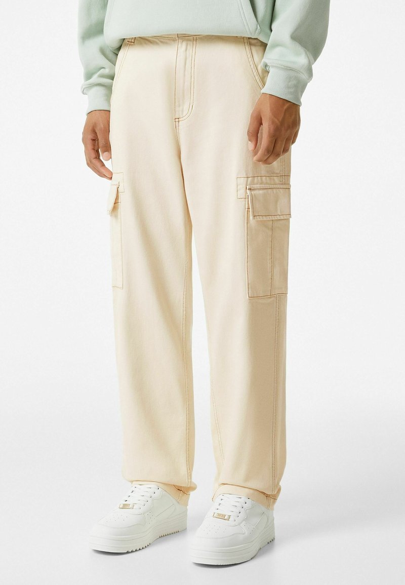 Pantalon cargo beige à coupe décontractée, avec poches latérales et coutures contrastées, associé à des baskets blanches sur un fond neutre.