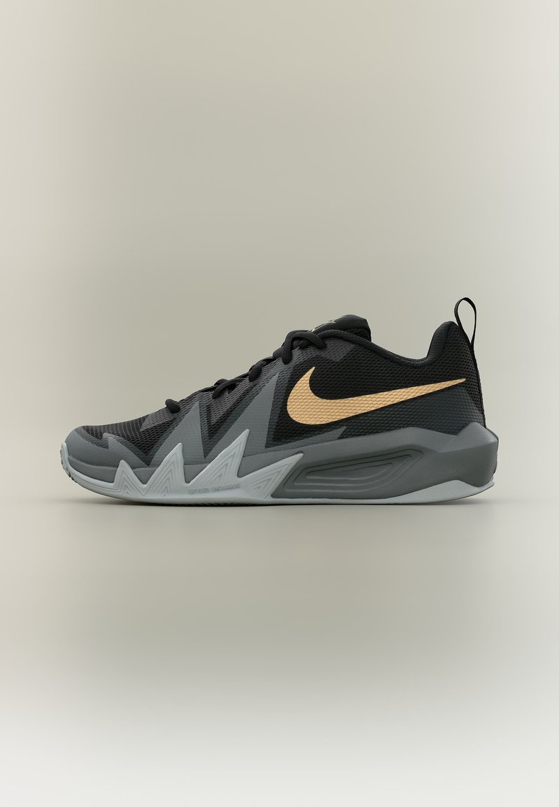 Μαύρο και γκρι χαμηλό sneaker της Nike με χρυσό swoosh, ζιγκ-ζαγκ σχέδιο στη σόλα και θηλιά στο τακούνι, εμφανίζεται σε πλευρικό προφίλ σε απλό φόντο.