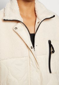 Quiltad krämfärgad jacka med en fluffig vit krage, svart dragkedjestängning och en sidoficka. Har en strukturerad, mjuk yta.