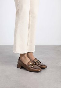 Zapatos de tacón bajo de cuero marrón con punta cuadrada, que presentan un detalle de hebilla dorada y una textura suave, combinados con pantalones de mezclilla crema.
