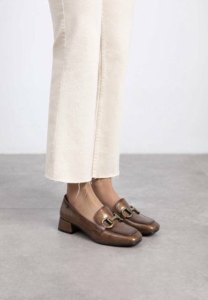 Zapatos de tacón bajo de cuero marrón con punta cuadrada, que presentan un detalle de hebilla dorada y una textura suave, combinados con pantalones de mezclilla crema.