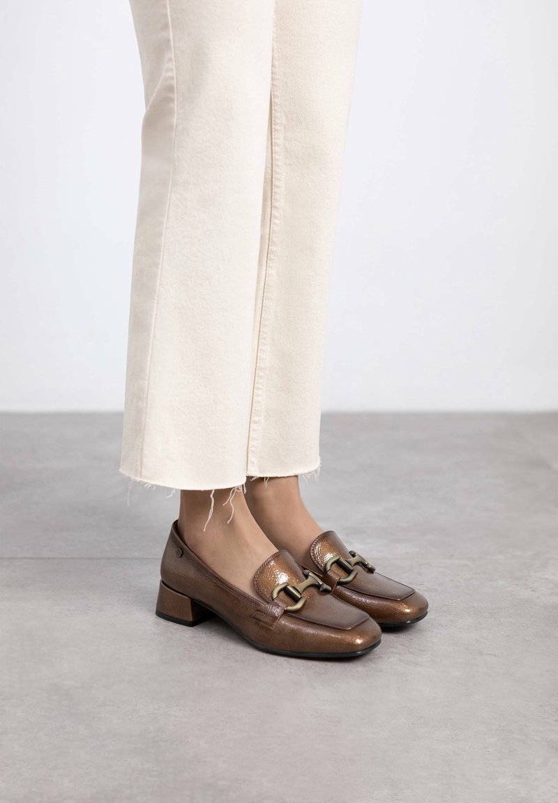 Zapatos de tacón bajo de cuero marrón con punta cuadrada, que presentan un detalle de hebilla dorada y una textura suave, combinados con pantalones de mezclilla crema.