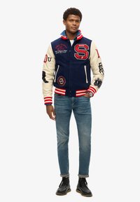 Chaqueta varsity en azul marino y crema con acentos rojos, que presenta parches bordados y rayas en los puños y el dobladillo. Combinada con jeans azules y botas negras.