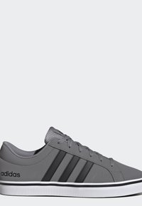 Zapatilla gris Adidas con tres franjas negras, diseño de cordones y suela blanca con detalle de línea negra, mostrada de lado.