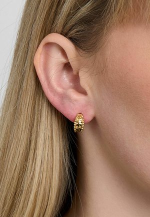 Közelkép egy fülről, amely hosszú szőke hajjal van, és apró, texturált arany karika fülbevalót visel.