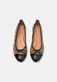 Paire de ballerines imprimé léopard avec bouts en cuir verni noir et petits nœuds noirs sur fond blanc.