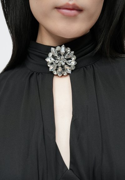 Fekete ruha magas nyakkal és kulcstartó kivágással, tiszta és ezüstszínű strasszokkal díszített kör alakú brossal.