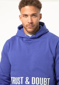 Sudadera azul hecha de una mezcla de algodón; cuenta con capucha ajustable, botones de presión y texto blanco que dice "CONFIANZA Y DUDAS" en la parte delantera.