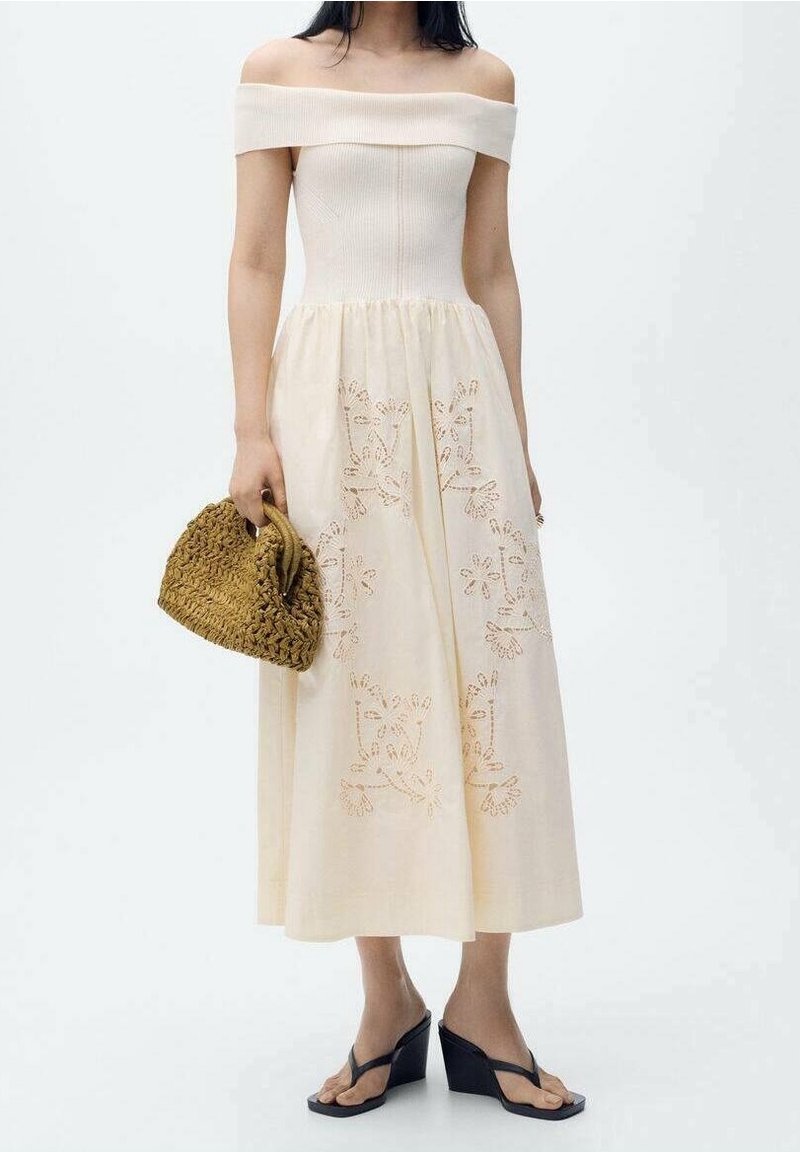 Femme portant une robe crème à épaules dénudées avec broderies florales, tenant un sac à main tissé et portant des sandales compensées noires.