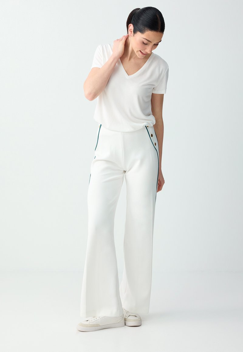 Femme en T-shirt blanc à col en V, pantalon blanc taille haute à jambes larges avec boutons latéraux, et baskets blanches, debout devant un fond uni.