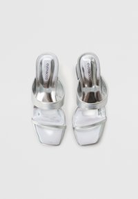 Sandales à talons argentées avec une finition métallique brillante. Présentent deux lanières sur le dessus et un design à bout carré. Texture douce et lisse.