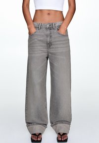 Jean en denim gris à taille haute et jambes larges, avec passants de ceinture, poches avant et ourlet effiloché. Porté avec des sandales noires.