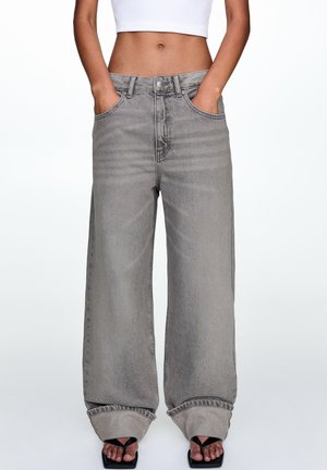 Jean en denim gris à taille haute et jambes larges, avec passants de ceinture, poches avant et ourlet effiloché. Porté avec des sandales noires.