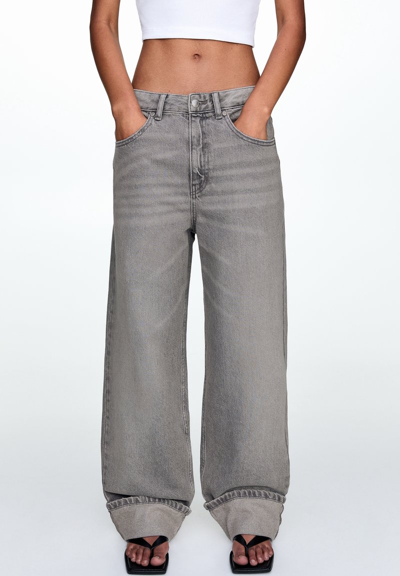 Jean en denim gris à taille haute et jambes larges, avec passants de ceinture, poches avant et ourlet effiloché. Porté avec des sandales noires.