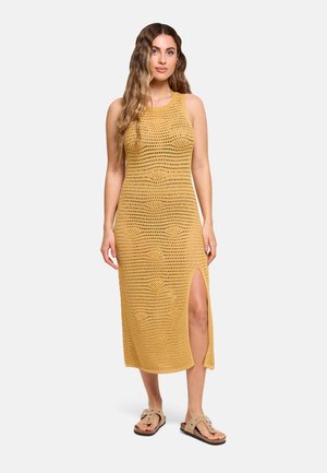 Mujer con cabello largo y ondulado lleva un vestido sin mangas de ganchillo amarillo mostaza con abertura lateral y sandalias beige, de pie sobre un fondo liso.